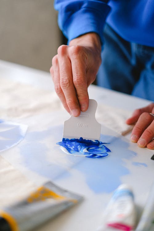 Unrecognizable male painter mixing blue and white paints while standing at table with tubes in art studio on blurred background