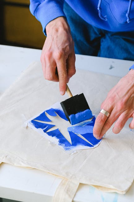 From above of unrecognizable male master using stencil and brush to draw sketch with blue paint on cloth bag while standing at table in studio