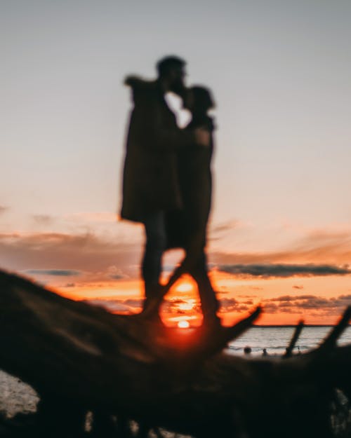 Free Defocused man cuddling and kissing unrecognizable woman in casual clothing while standing on coast of ocean against vibrant sky covered with orange clouds Stock Photo
