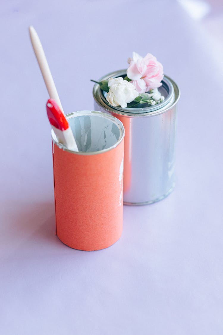 Paint Cans, Artificial Flowers And Paint Brushes On Lilac Surface