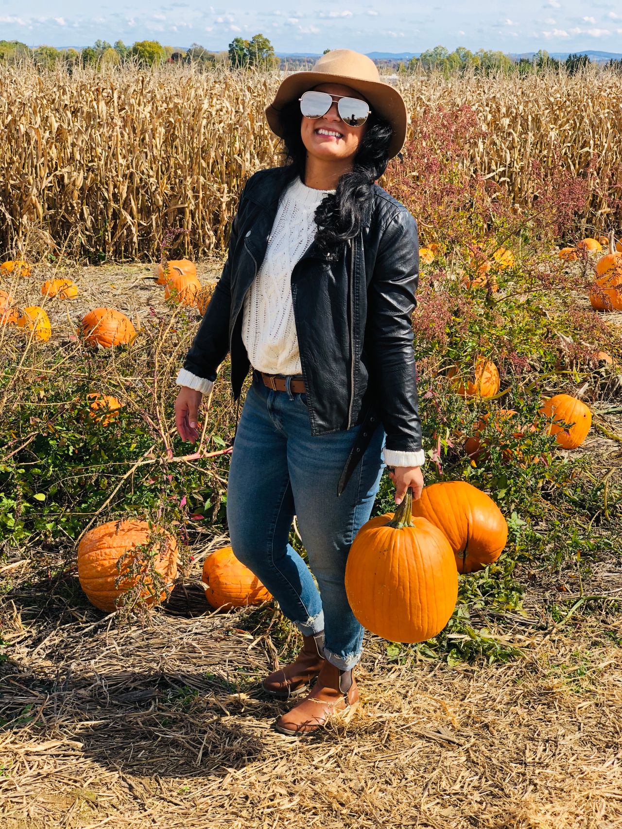 Woman in a black leather jacket over a cream cable-knit sweater and jeans holding a pumpkin in a cornfield patch