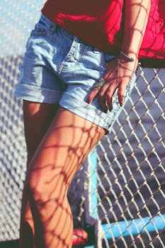 Woman in denim shorts with jewelry beside a chain link fence casting shadows.