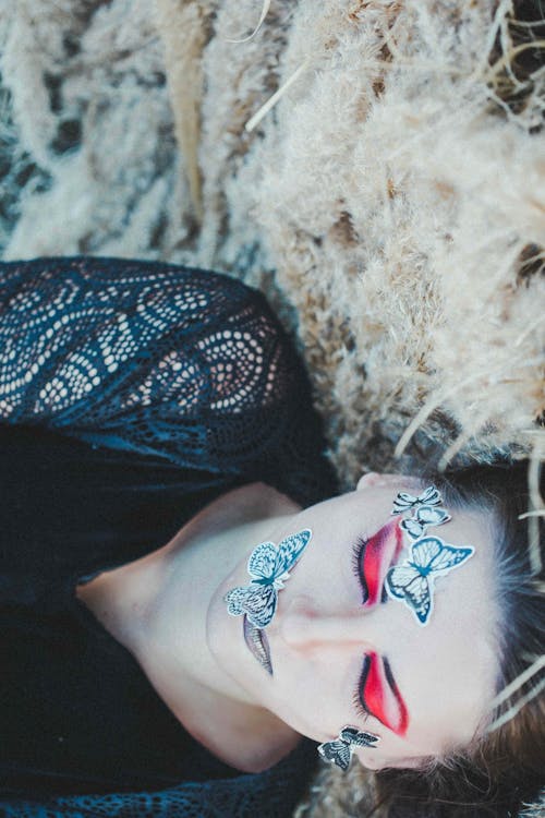 Top view of young trendy female with colorful eye shadow on closed eyes and ornamental butterflies on face resting on fluffy plants