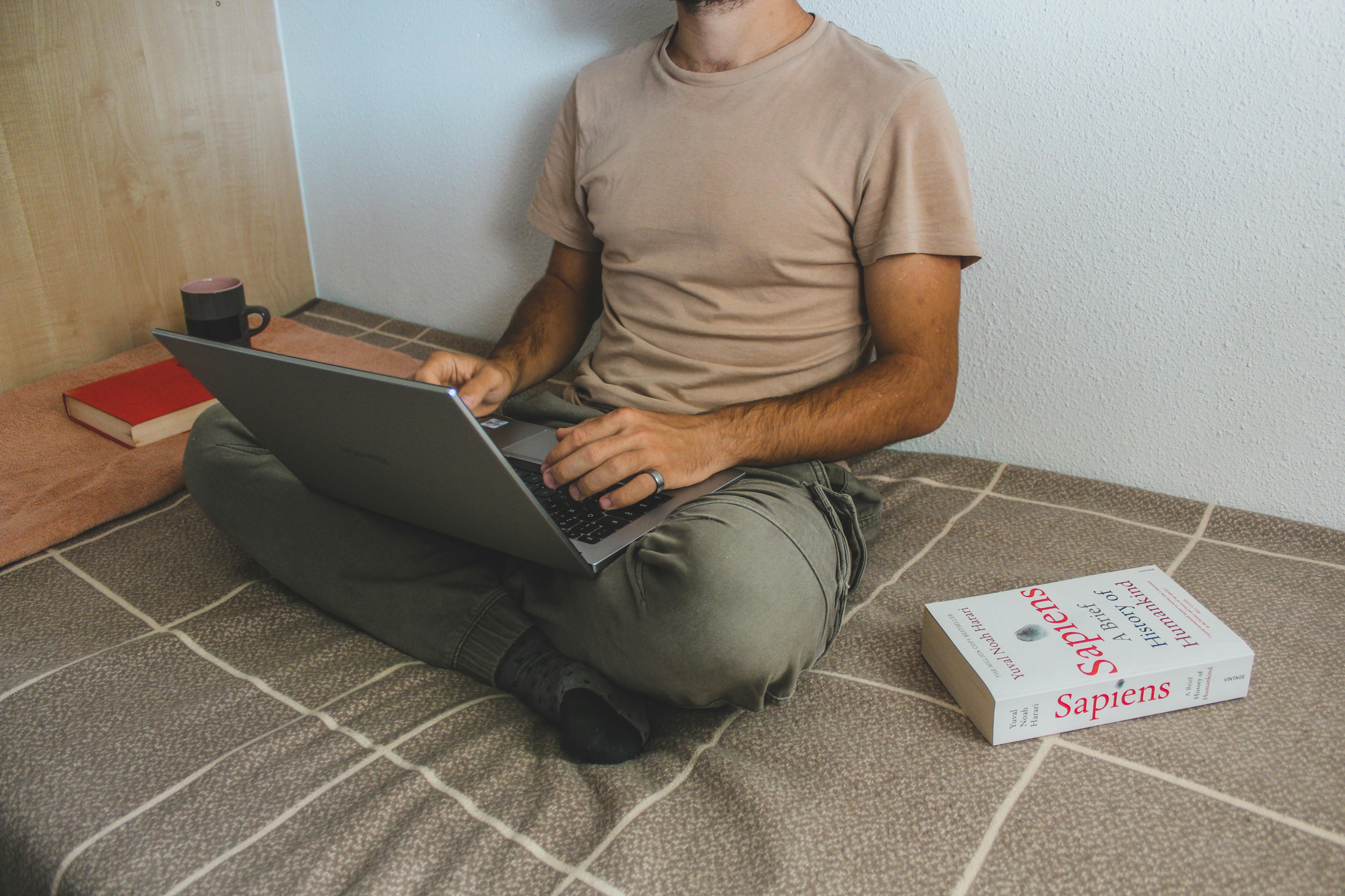 Free Man working remotely on a laptop, surrounded by books in a cozy indoor setting, reflects modern digital lifestyle. Stock Photo