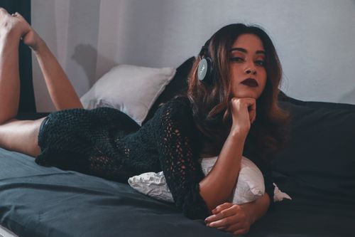 Free Woman lying on bed wearing headphones and black dress, creating a relaxed and stylish atmosphere. Stock Photo