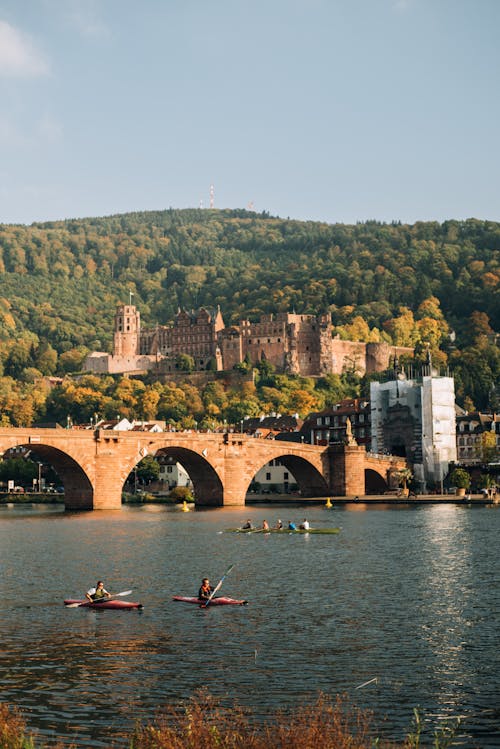 Picturesque view of classic stone castle in forest on mountain and brick bridge over calm rippling river with boats