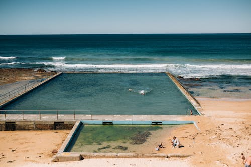 Sandy seashore with swimming pool near waving water with foam under clear blue sky