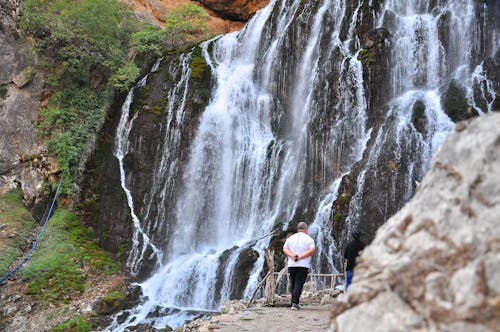 Free Back view of unrecognizable tourists in casual clothes walking on walkway while admiring waterfall in mountainous area Stock Photo