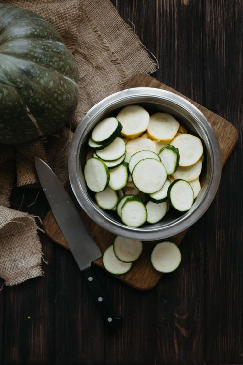 Free High-angle shot of sliced zucchini in a stainless bowl with a knife on a wooden board, perfect for food preparation. Stock Photo