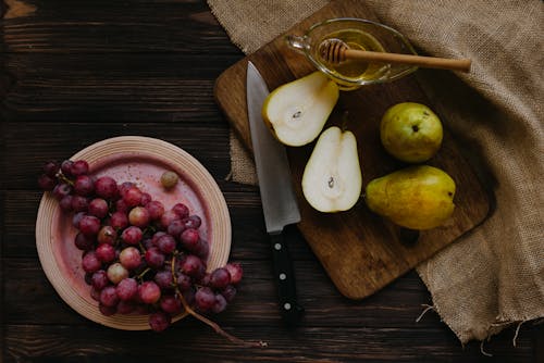 Top view composition of cut pears with honey in bowl on cutting board and plate with grapes on wooden rustic table