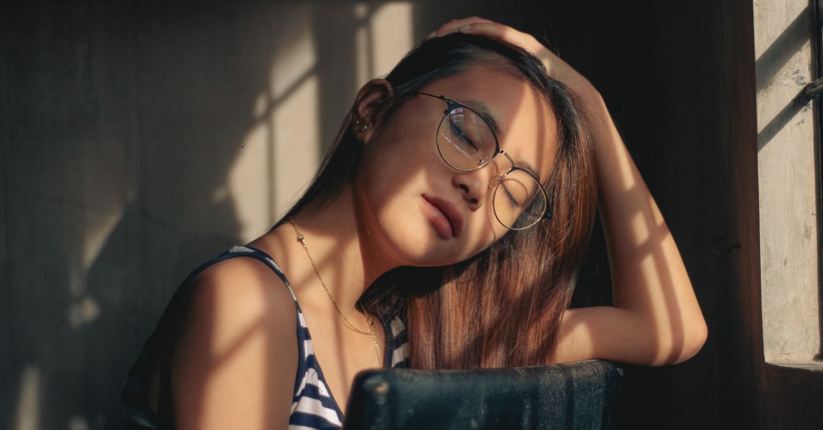 Dreamy Asian teen girl with closes eyes adjusting hair sitting on chair near window in sunny day