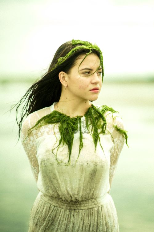 Free Thoughtful young ethnic female in dirty white dress and see weed on head and chest looking away with arms behind back in daytime on blurred background Stock Photo