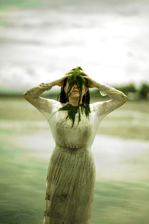 Faceless woman with see weed on face by pond in daytime