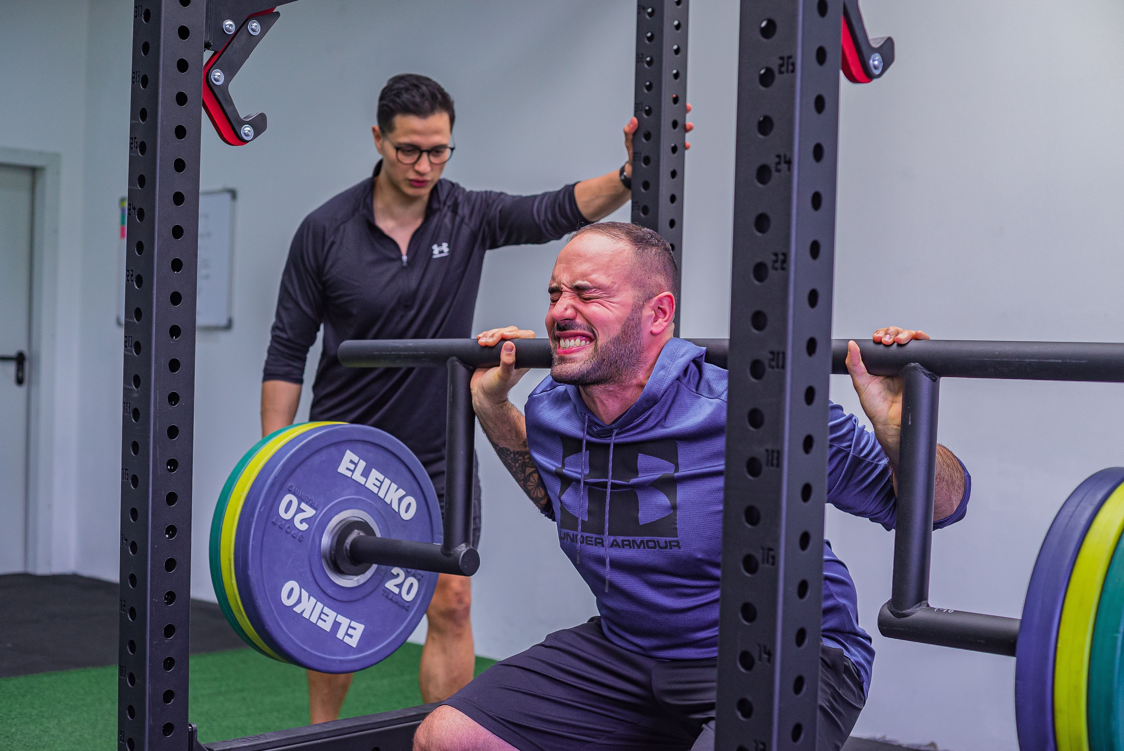 Muscular sportsman in activewear squatting with heavy barbell with effort while listening trainer in gym