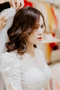 Beautiful Asian bride in white gown having her veil adjusted by a stylist indoors.