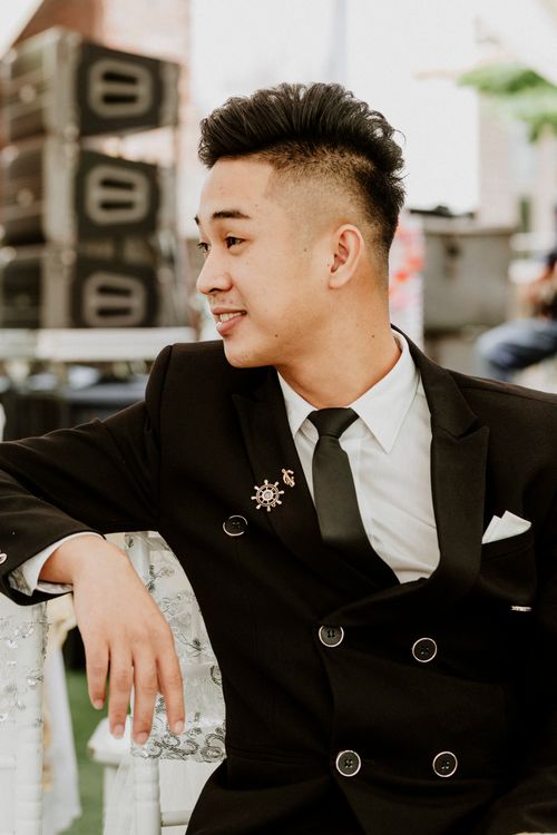 Free Portrait of a young man in a black suit sitting at an outdoor event, smiling and looking sideways. Stock Photo