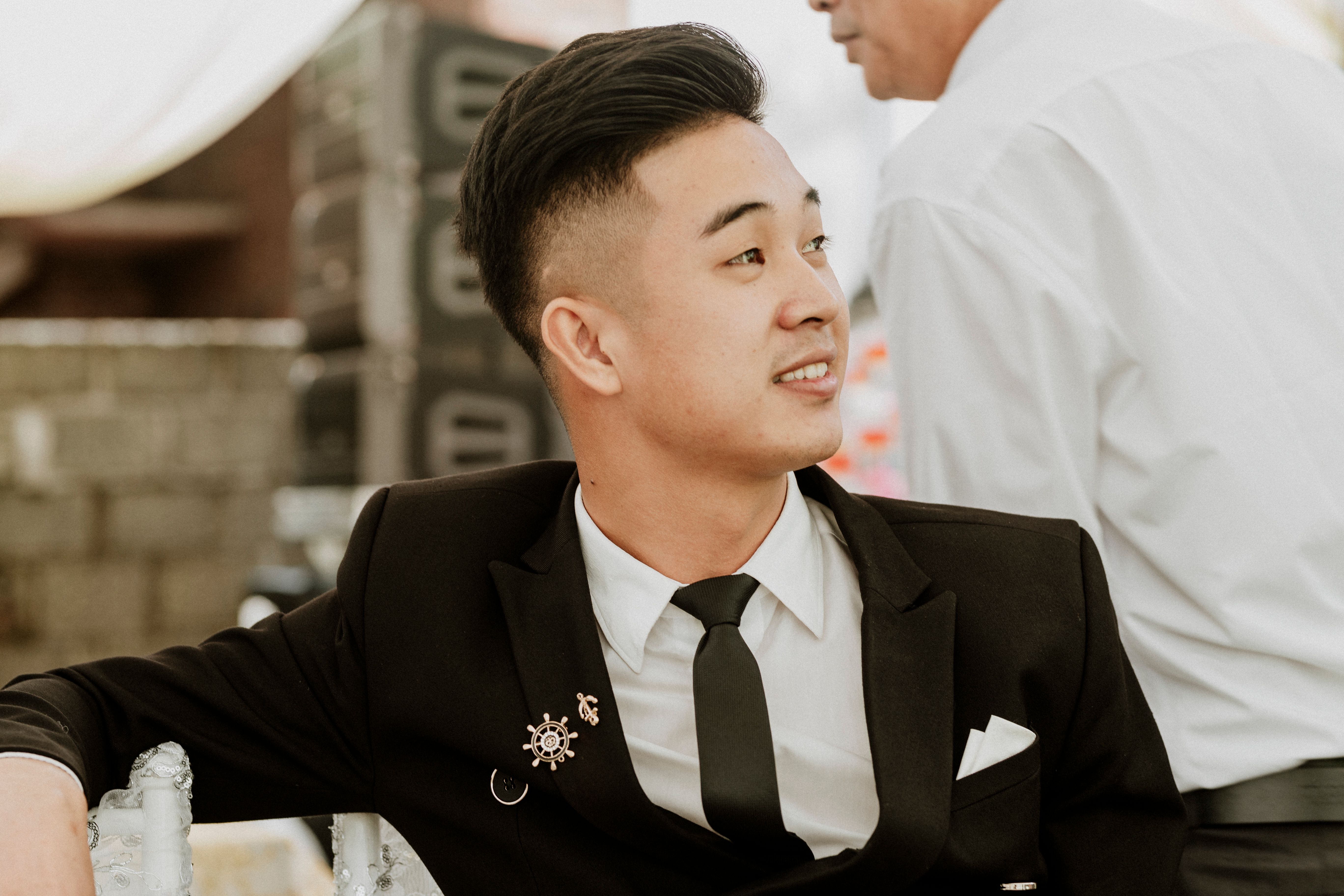 Asian man in black suit and tie looking away, seated outdoors against a soft background.