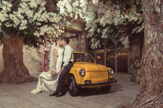 Full body of happy bride and groom kissing in yard with decorated trees near yellow retro car