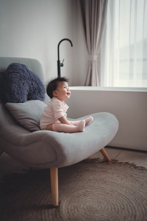 Free Side view full body joyful Asian toddler in white outfit sitting on comfy chair in light living room and laughing Stock Photo