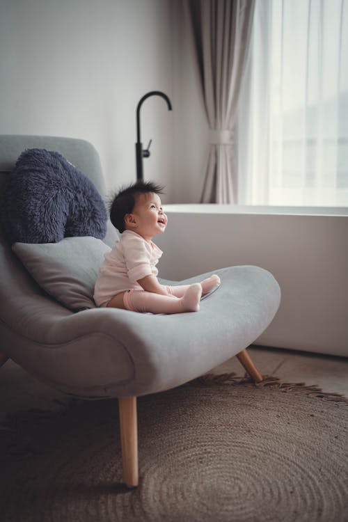Side view full body joyful Asian toddler in white outfit sitting on comfy chair in light living room and laughing
