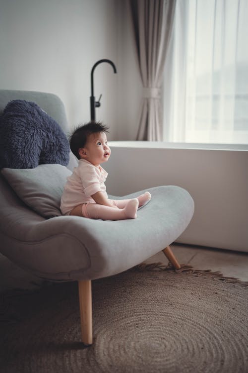 Free Side view full length playful Asian toddler wearing white cozy clothes sitting on soft chair at home and looking away with interest Stock Photo