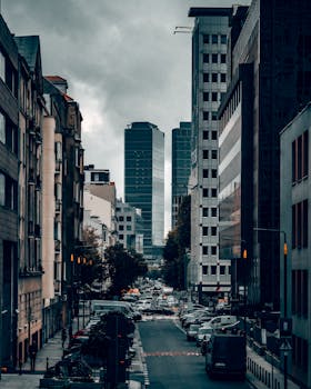 A moody street scene in Warsaw, Poland with tall skyscrapers and parked cars.