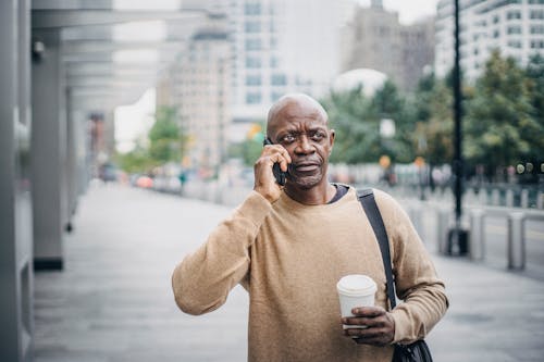 Focused adult African American businessman with coffee cup walking in downtown and talking on phone