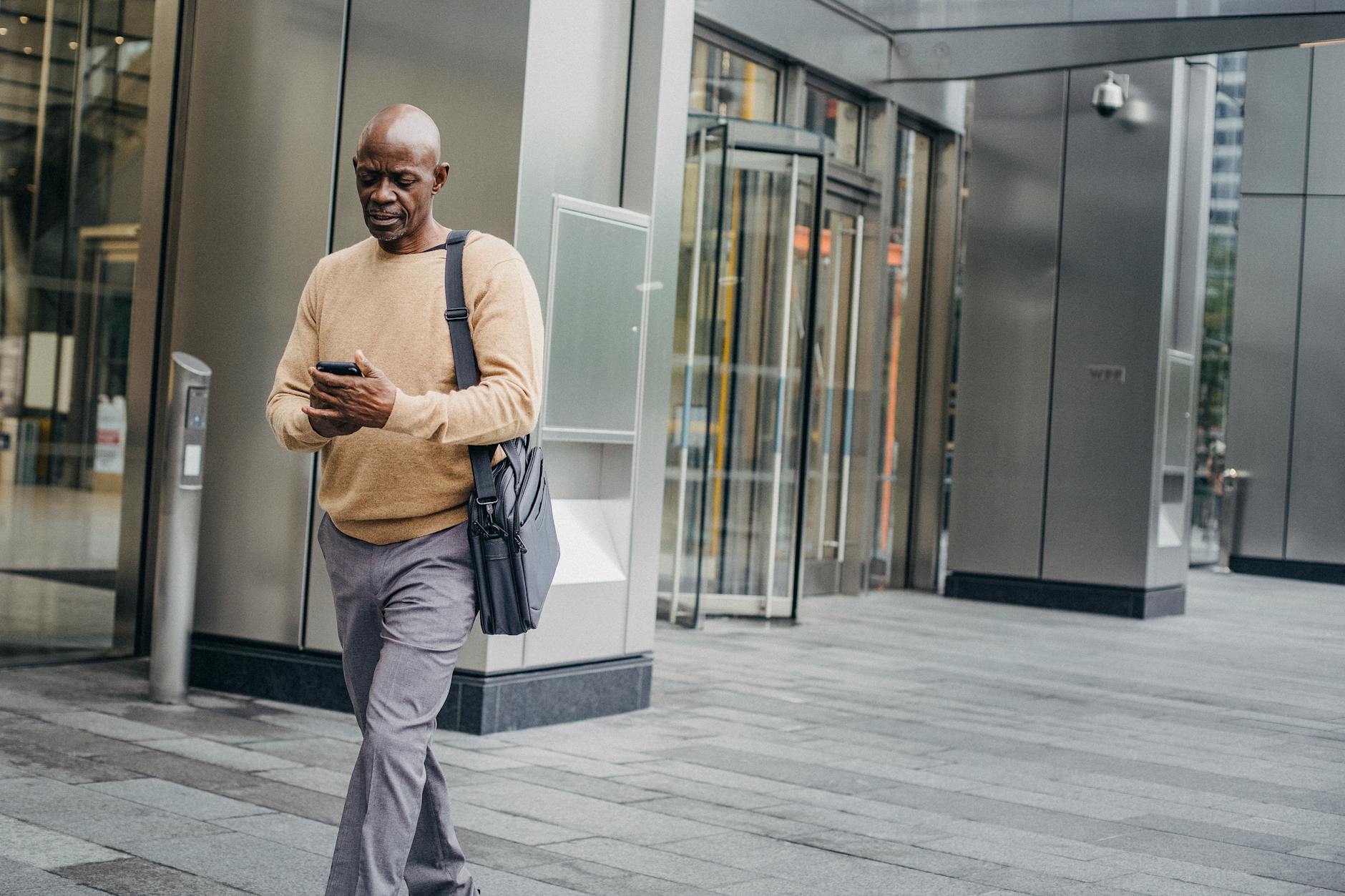 Mature bald black man in elegant clothes and with case bag texting on smartphone while walking down street