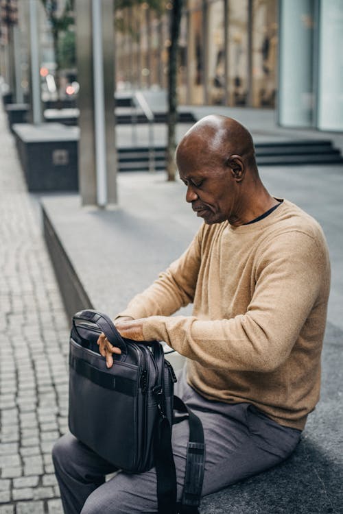 Focused bald ethnic businessman with case bag on knees sitting on fence in city street