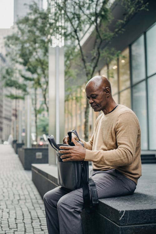 Modern African American male worker with laptop in case bag sitting on street in downtown