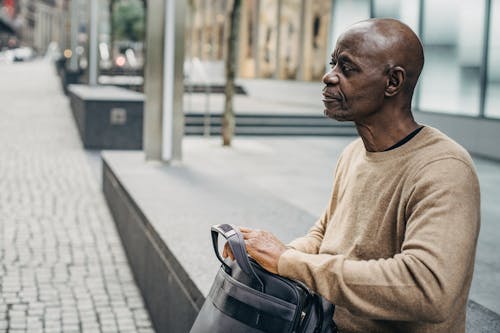 Mature black man sitting on street fence with bag and looking away pensively