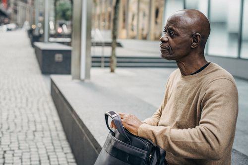 Free Mature black man sitting on street fence with bag and looking away pensively Stock Photo