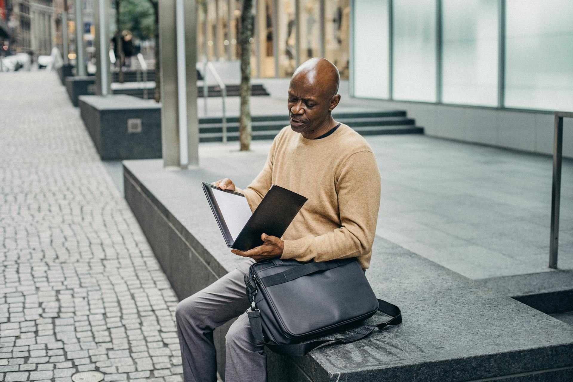 Mature African American male opening folder with document while sitting on marble fence in downtown