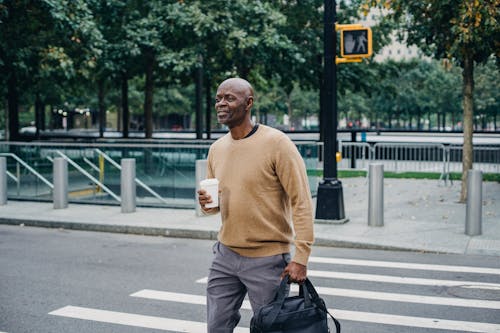 Free Mature African American man carrying disposable coffee cup and personal bag crossing street in downtown Stock Photo