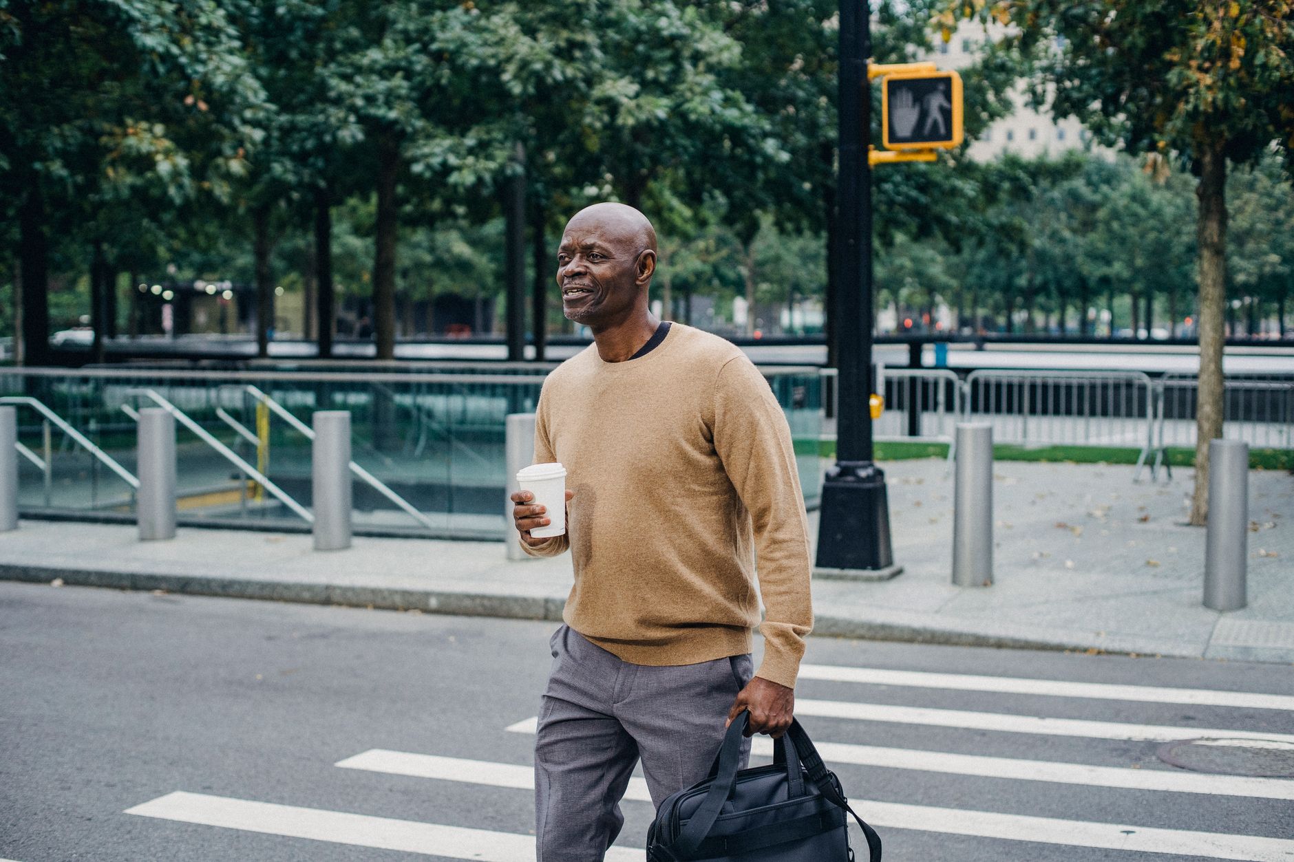 Mature African American man carrying disposable coffee cup and personal bag crossing street in downtown