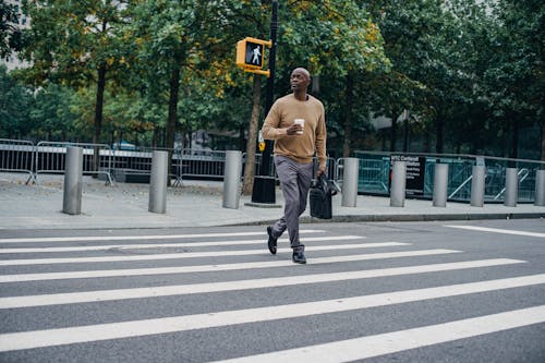 Full body of African American male walking on pedestrian crossing on asphalt road with cup of hot drink and briefcase