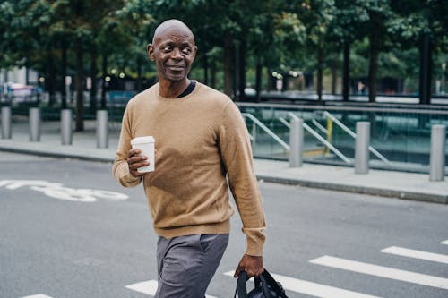 Cheerful middle aged African American male with cup of hot drink and briefcase walking on pedestrian crossing on street while looking away