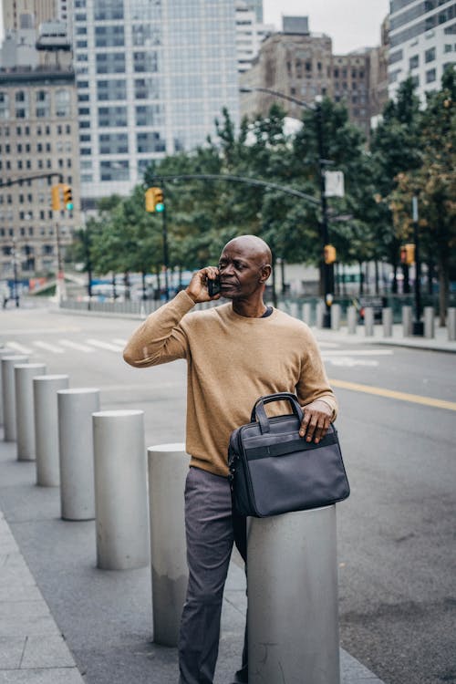 Serious African American bold male talking on smartphone while standing near stone columns on roadside with briefcase on street in city