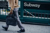 Side view of unrecognizable African American worker walking along green fence near entrance of subway station with briefcase in hand