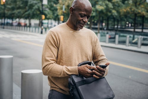 Content middle aged African American male with leather bag surfing cellphone while standing near posts on roadside with blurred street