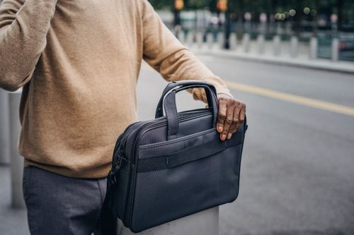 Free Unrecognizable African American male wearing brown sweater standing on roadside with black bag on stone pillar near blurred road in street Stock Photo