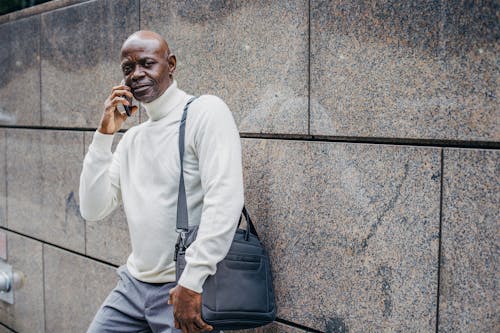 Serious middle aged African American male talking on smartphone and looking at camera while standing with bag on shoulder near marble wall