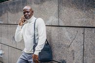 一位表情严肃的中年非裔美国男性扛着包站在大理石墙边，一边用手机通话，一边看向镜头