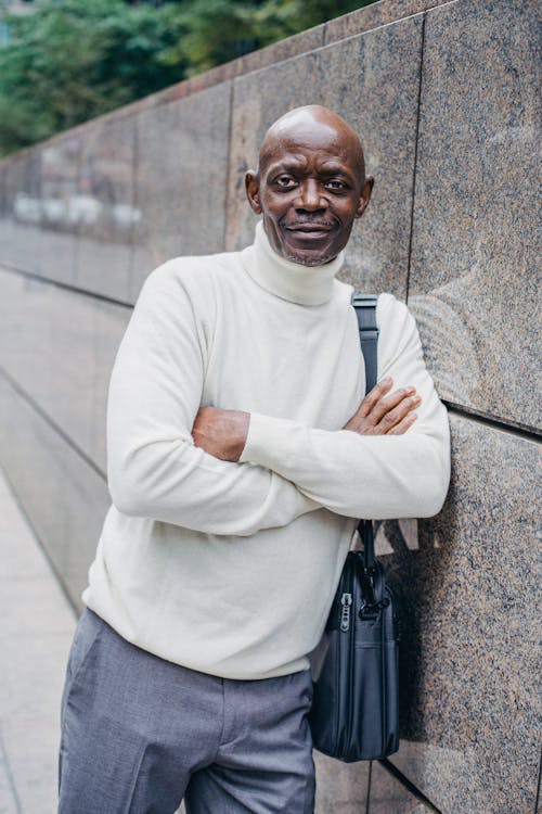 Content middle aged African American male wearing white sweater with bag on shoulder leaning on barrier while standing on street with crossed arms and looking at camera