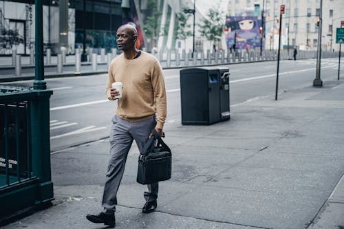 Full body of pensive African American male walking on pavement near asphalt road with takeaway coffee and black handbag in hand