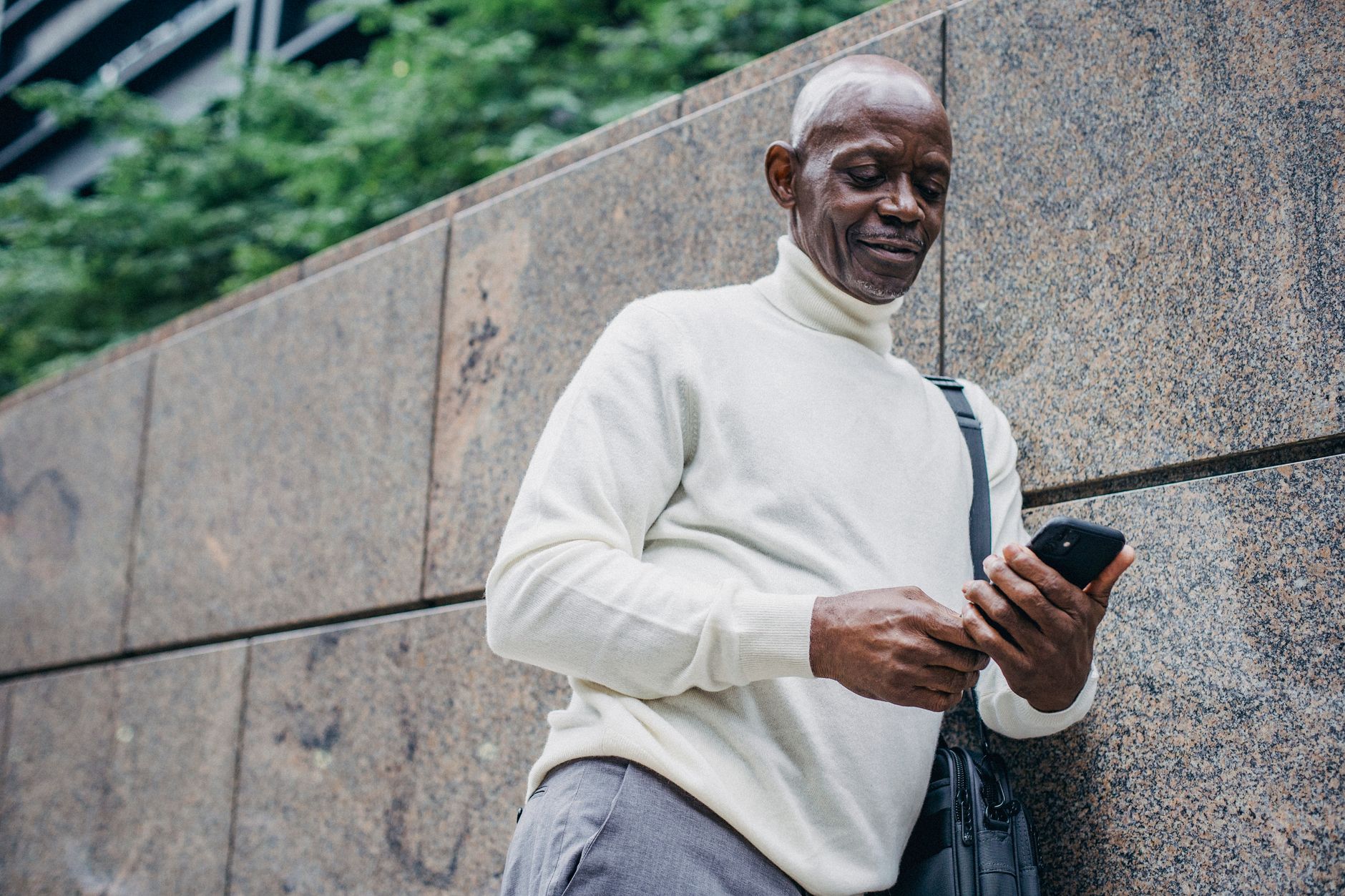 Low angle of cheerful African American male in trendy outfit browsing smartphone while leaning on wall