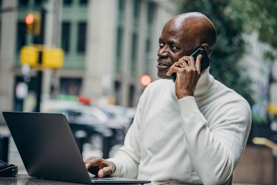 African American man in casual clothes sitting at table with laptop and talking on smartphone during