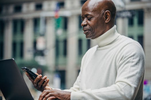 Side view of concentrated African American businessman in casual clothes browsing smartphone while working on laptop on city street