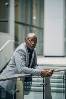一位自信的非裔美国商人穿着正式西装，靠在玻璃栏杆上，拿着手机，目光移向别处