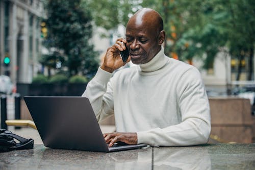 Concentrated African American man talking on mobile phone while working on laptop on city street